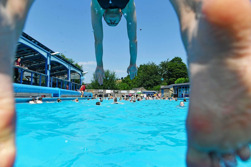 Looking under a person, you see them dive into an azure pool with the feet close to the camera's lens.