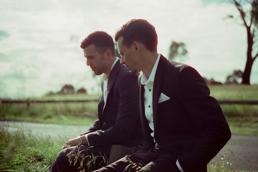 Two young men with dark hair wearing black suits with white shirts underneath sit at a roadside bench surrounded by grass.