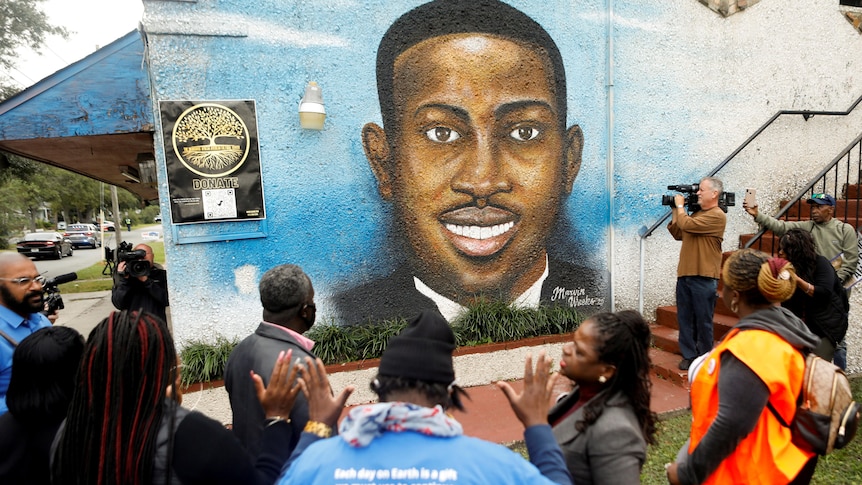 People gather at a wall with a mural painted of Ahmaud Arbery. 