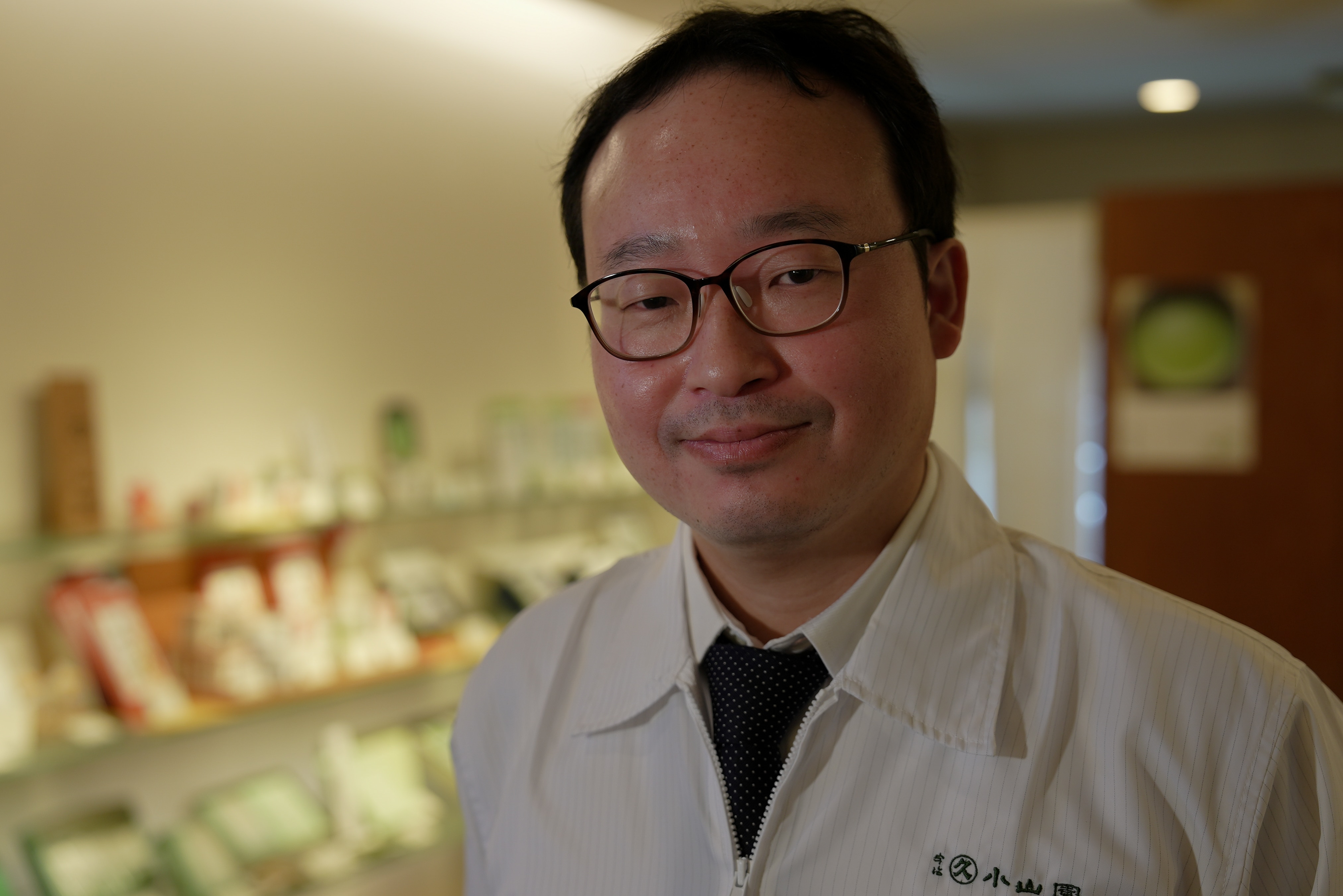 Um japonês de óculos e jaleco sorri em uma sala.