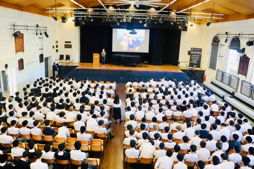 A school assembly with a presentation at the front. There are rows of students sitting down in white school shirts.