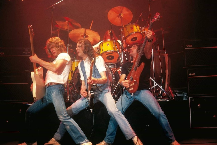 Three middle-aged guitarits wearing denim play their instruments at the front of a brightly lit stage.