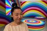Japanese-Australian women stands in front of a room with walls, furniture and Tv screen covered in giant rainbows 
