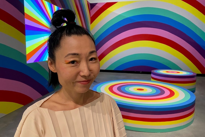 Japanese-Australian women stands in front of a room with walls, furniture and Tv screen covered in giant rainbows 