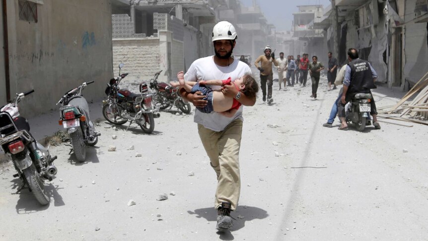 A man in a white helmet carries a young girl in a red T-shirt along a rubble-strewn street