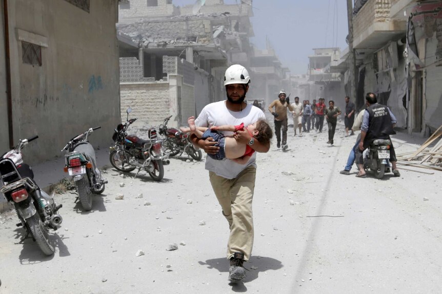 A man in a white helmet carries a young girl in a red T-shirt along a rubble-strewn street