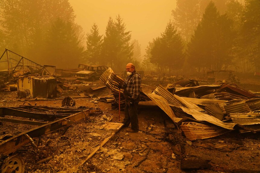 A man stands in the wreckage of his fire destroyed home with an orange sky.