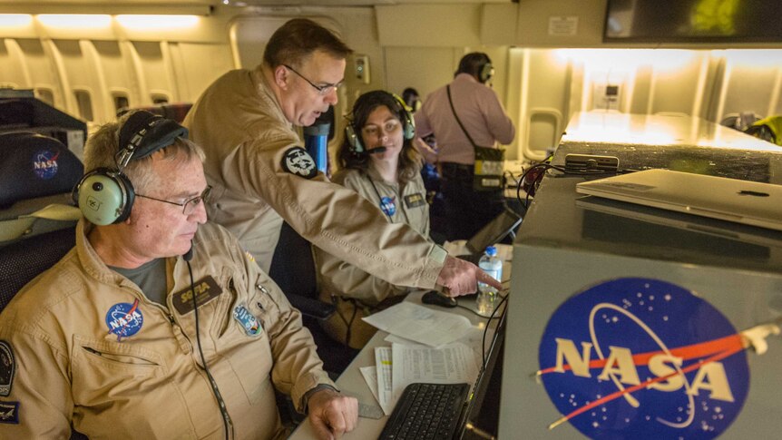 Two people wearing NASA jumpsuits sit at workstations while a man leans past them pointing at one of the screens.