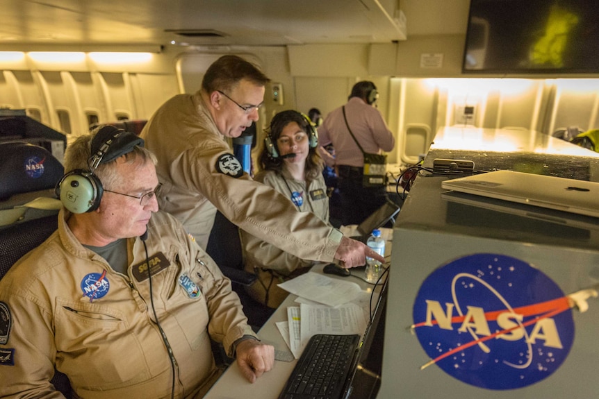 Two people wearing NASA jumpsuits sit at workstations while a man leans past them pointing at one of the screens.