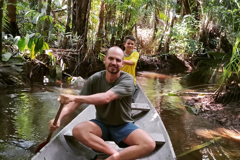 A man and a boy paddle in shallow water surrounded by rainforest.