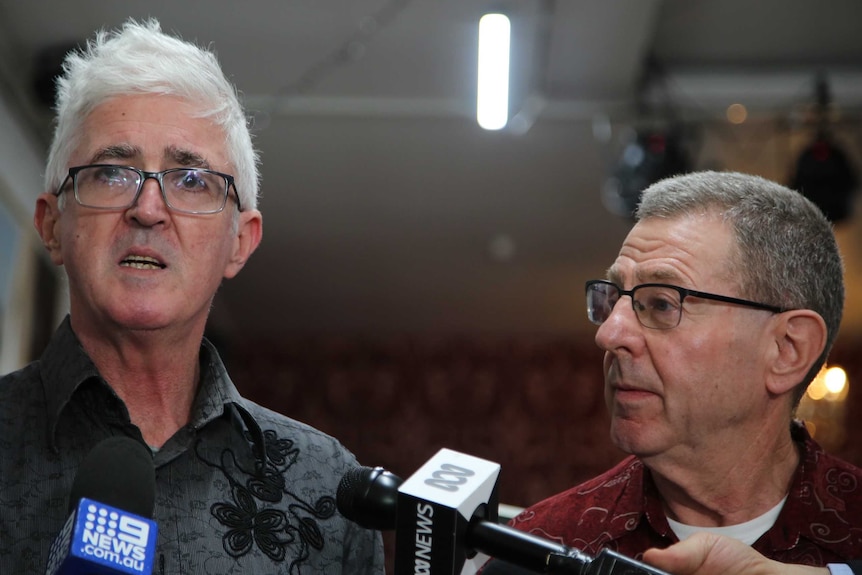 A man with white hair speaks into microphones, while a man with grey hair looks on.