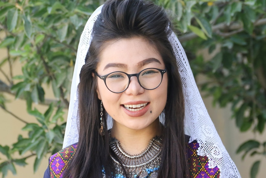 A woman with black hair and wearing a white hijab and colourful dress smiles at the camera.