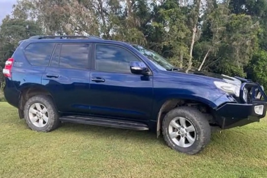 A dark-coloured four-wheel drive parked on some grass in front of a forested area.