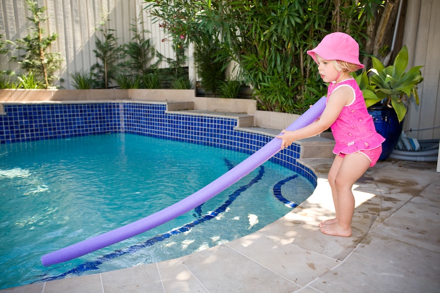 Child playing with a pool noodle beside backyard pool
