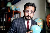 Sami Shah dressed in a suit holding a globe of the world.