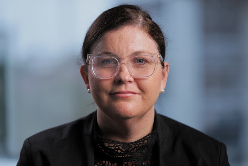 Portrait of a woman with dark hair, clear framed glasses and wearing a black blazer.