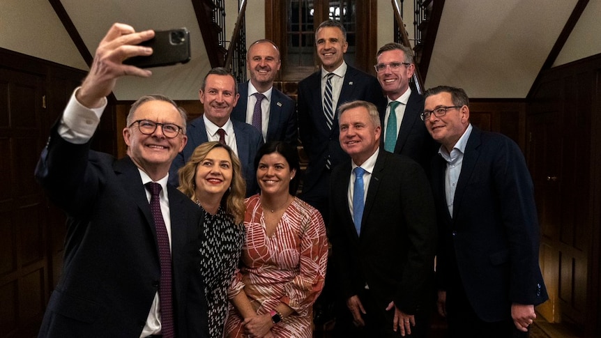 Albanese holds a phone up, smiling. The state leaders crowd behind him, also smiling.