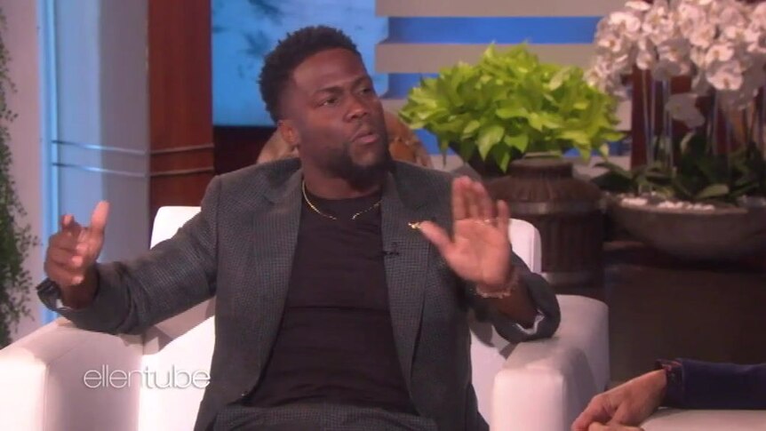 Kevin Hart sitting on a chair in a TV studio, arms outstretched while making conversational gestures.