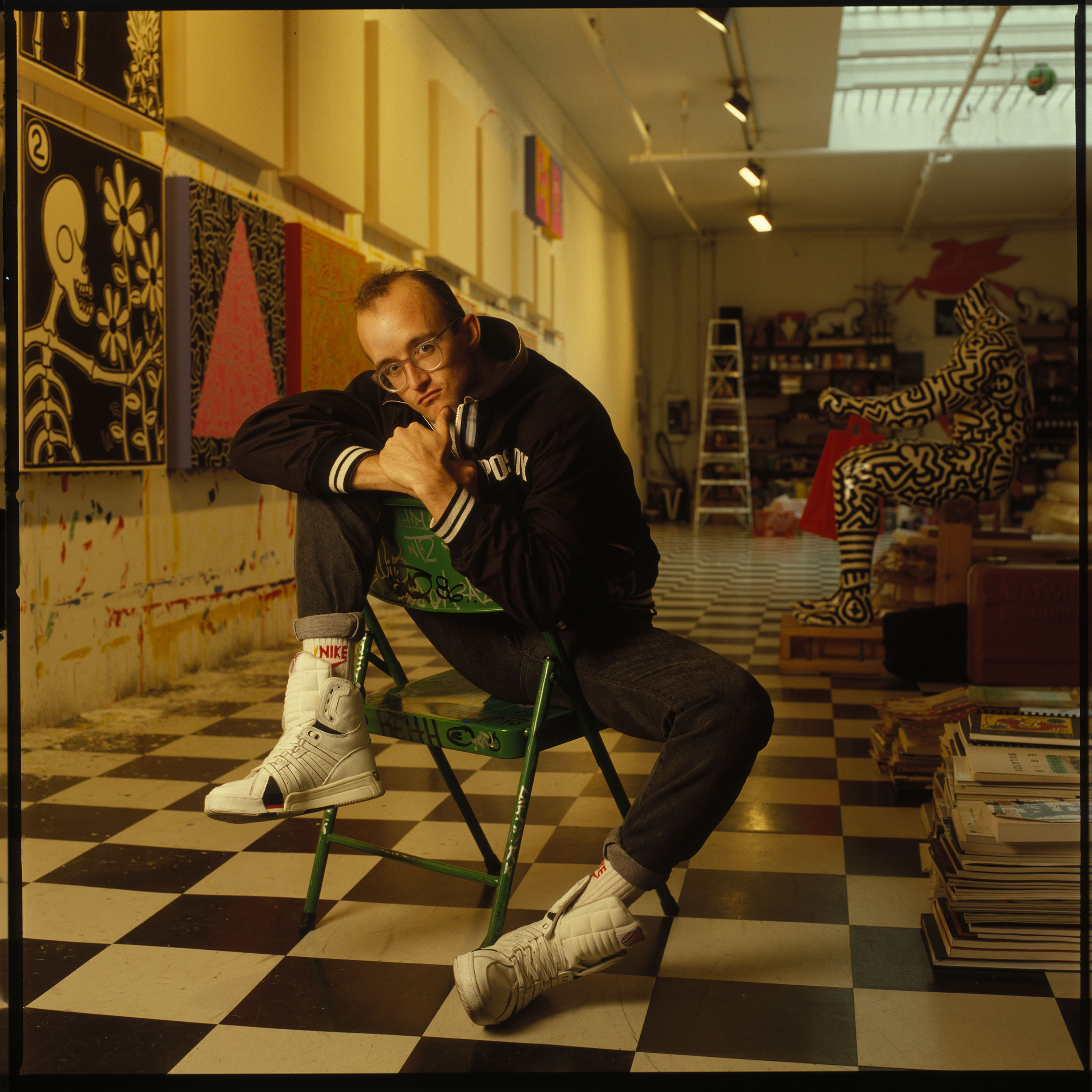 Square colour photograph of artist Keith Haring sitting on green folding chair in his chequered floor artist studio.