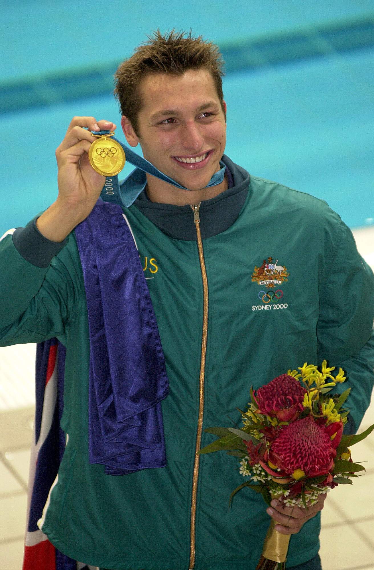 A male Australian swimmer smiles as he holds up a gold medal with his right hand after winning at the 2000 Sydney Olympics.