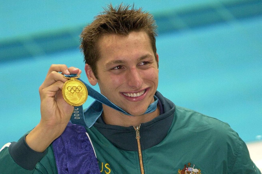 A male Australian swimmer smiles as he holds up a gold medal with his right hand after winning at the 2000 Sydney Olympics.