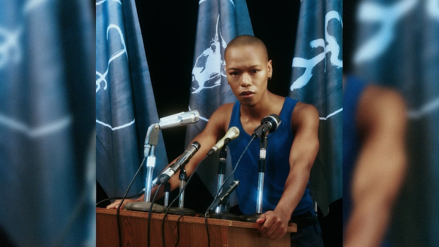 Nakhane stands at a podium about to speak into some microphones. There are blue flags behind them.