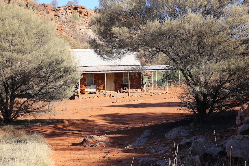 A small cottage in the outback of the Northern Territory is surrounded by rocky escarpment and bush trees. 