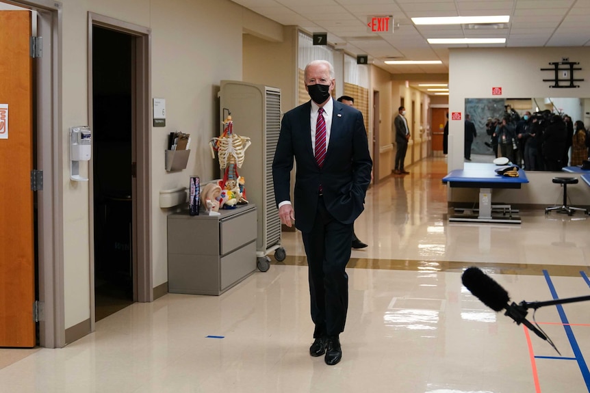 A tall, elderly man in dark suit, white shirt and tie and face mask walks along a hospital corridor.
