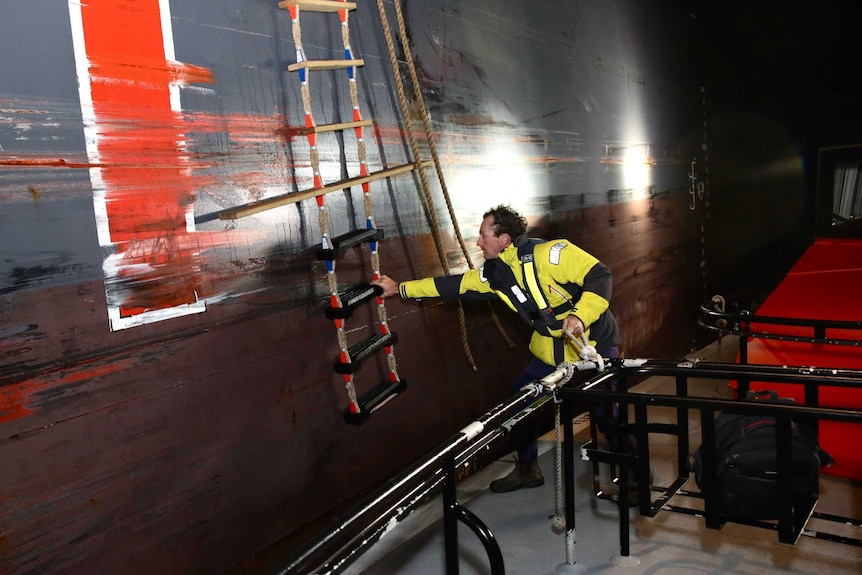 A man stands on a small boat next to a tanker ship with a rope ladder on the side.