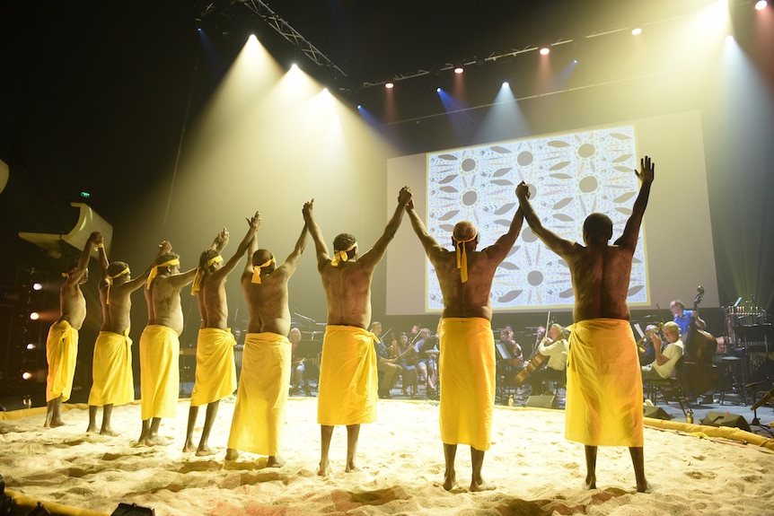Line of 8 men with ochred torsos standing on sand circle on stage holding hands in air, facing artwork projection.