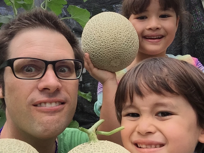 Australian journalist Scott McIntytre, wearing glasses, looks at the camera with his two children. They are holding rockmelons.