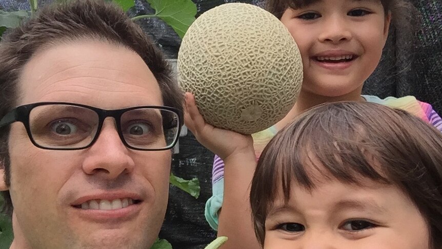 Australian journalist Scott McIntytre, wearing glasses, looks at the camera with his two children. They are holding rockmelons.