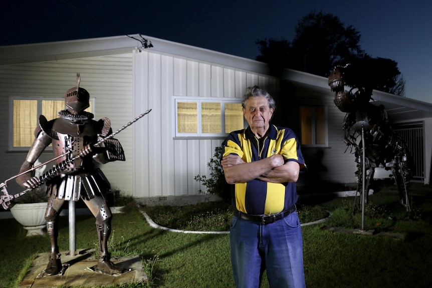 A man stands at night time in his front yard in front of two large recycled metal sculptures