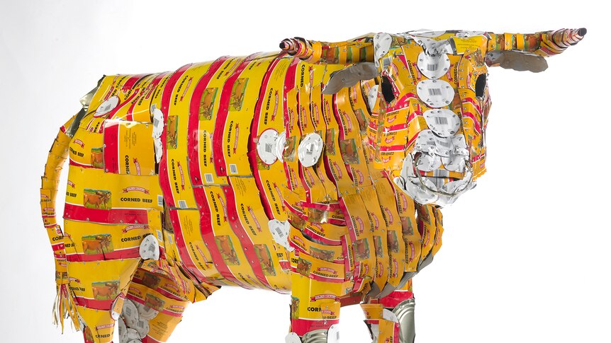 A bull art sculpture made out of flattened corned beef cans stands infront of a white background.