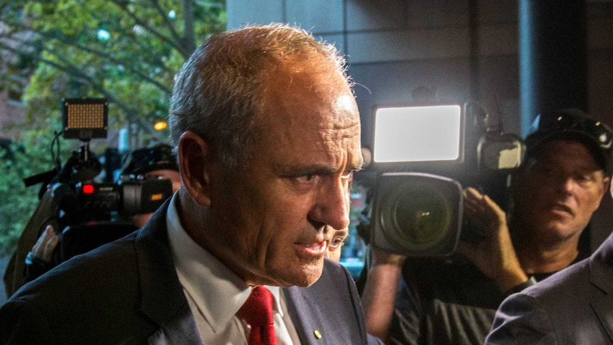 Ken Henry looks concerned as a 7 News journalist puts a microphone in front of his face, with cameras filming in the background.