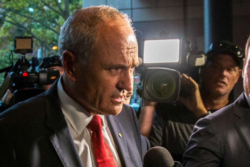 Ken Henry looks concerned as a 7 News journalist puts a microphone in front of his face, with cameras filming in the background.