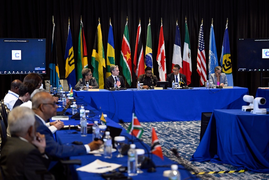 People sit at long tables with blue tablecloths and world flags in the background.