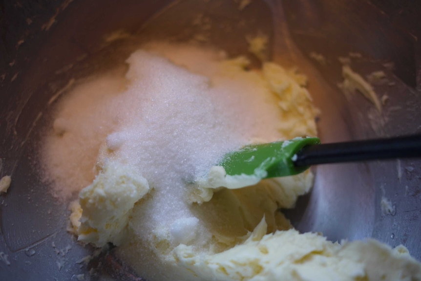 Butter with white sugar on top are in a silver mixing bowl and there's a green spatula in it ready to cream them together.