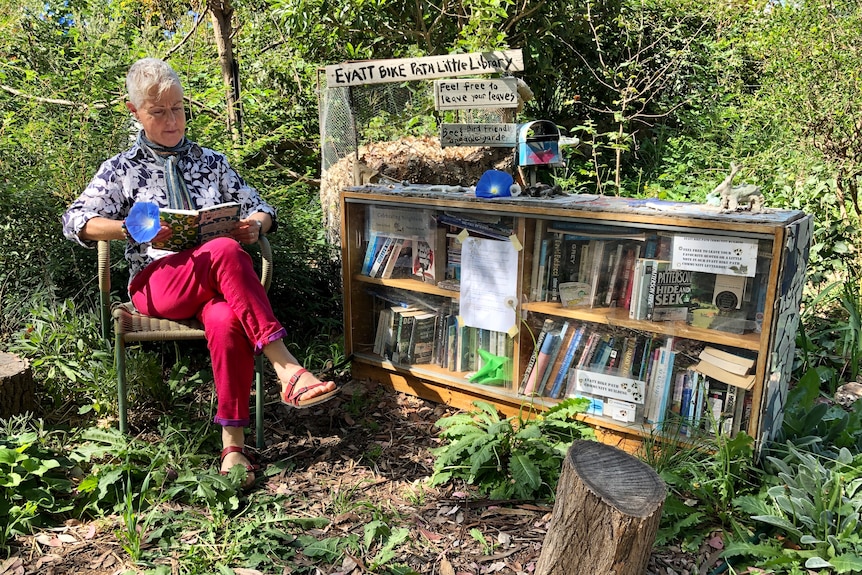 A woman sits outside in the sun on a chair next to a bookshelf of books.