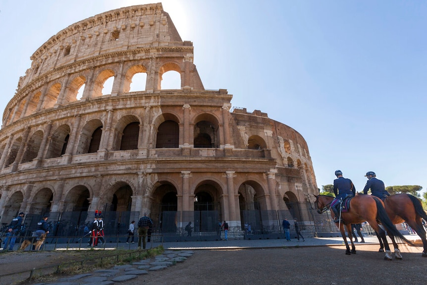 You look up at Rome's Colosseum on a bright day, with horse-mounted police and men in Carabinieri uniform patrolling.