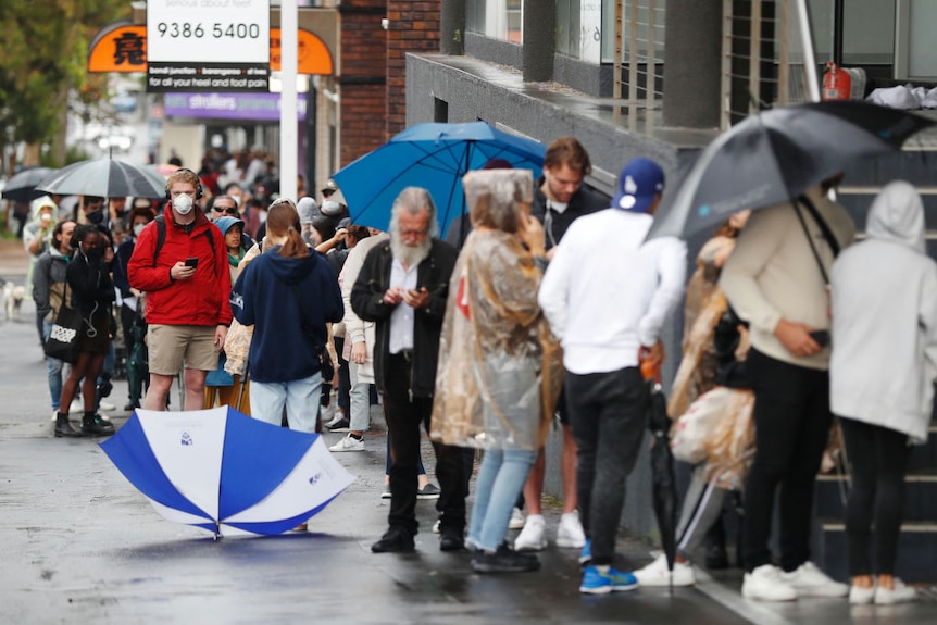 A long queue outside a Centrelink office extends down a street with people wearing masks and carrying umbrellas. Mood is tense.