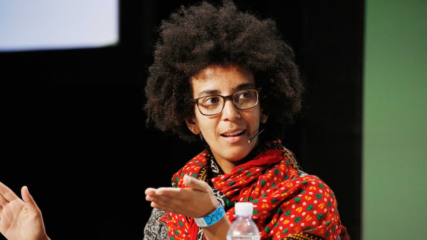 Black woman with glasses, brown hair, glasses and a red and green patterned scarf. She is speaking with a headset microphone on