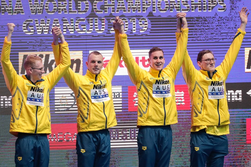 Four male swimmers wearing green and gold track suits raise their arms on a medial podium.