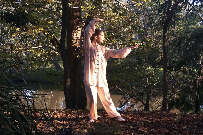 A woman moves her arms slowly above her head while standing in amongst trees.