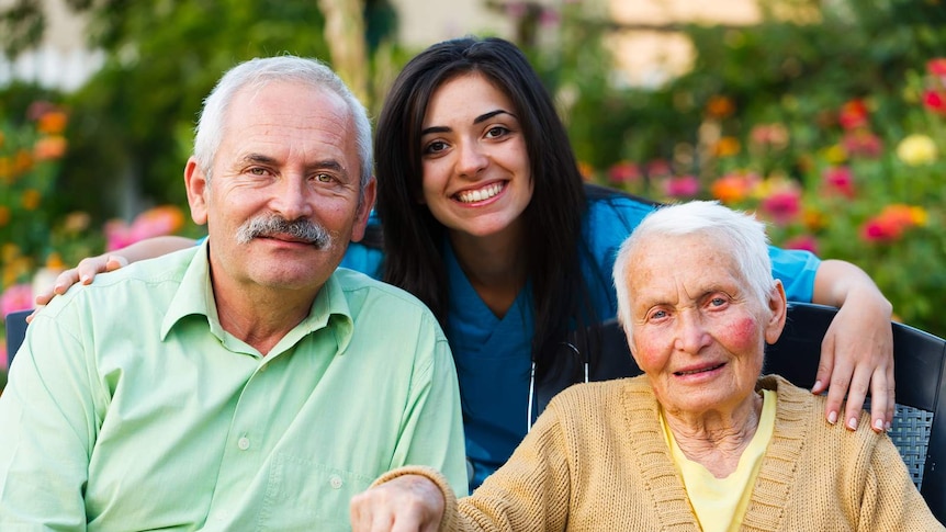 A young woman with dark hair is smiling with her arms around an elderly man and an elderly woman