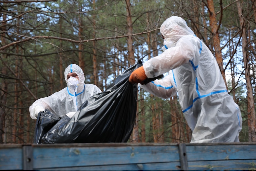 Two men in protective clothing hold a body bag on the back of a truck.