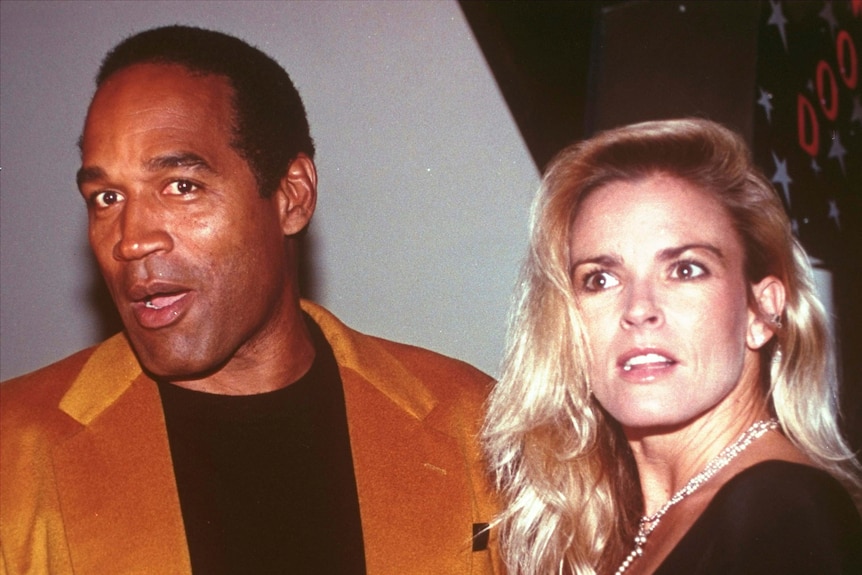 OJ Simpson, wearing a gold-orange blazer and black t-shirt, stands next to Nicole Brown Simpson, wearing a black top.