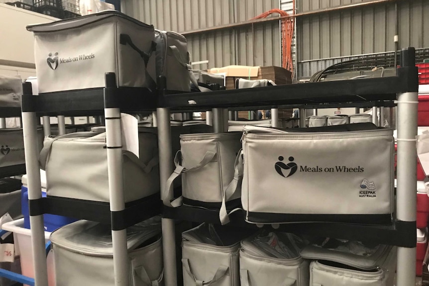 Grey cool bags with the Meals on Wheels logo sit on trolleys in an industrial kitchen.