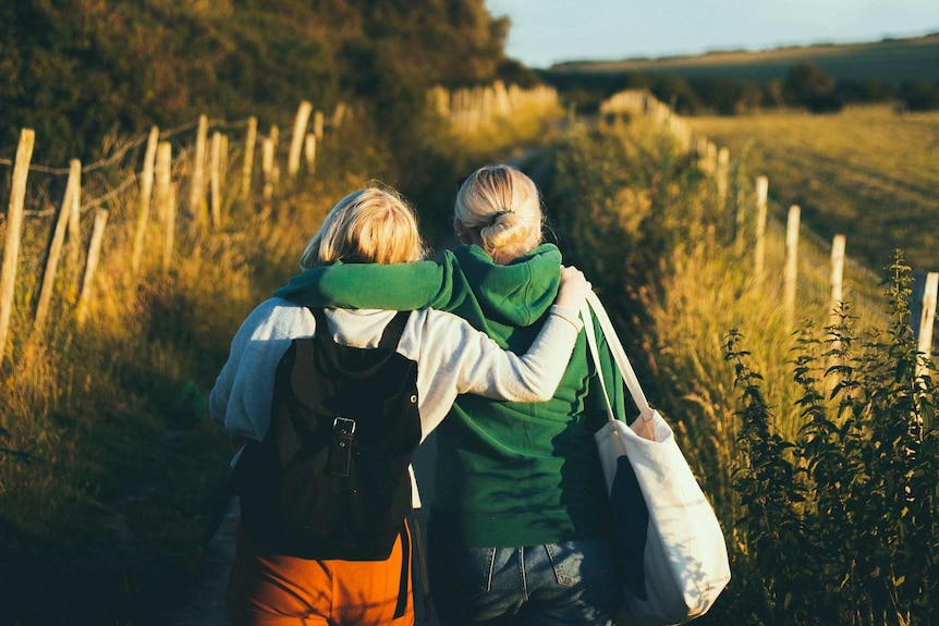 Two women walk through a field with their arms around the other's shoulders