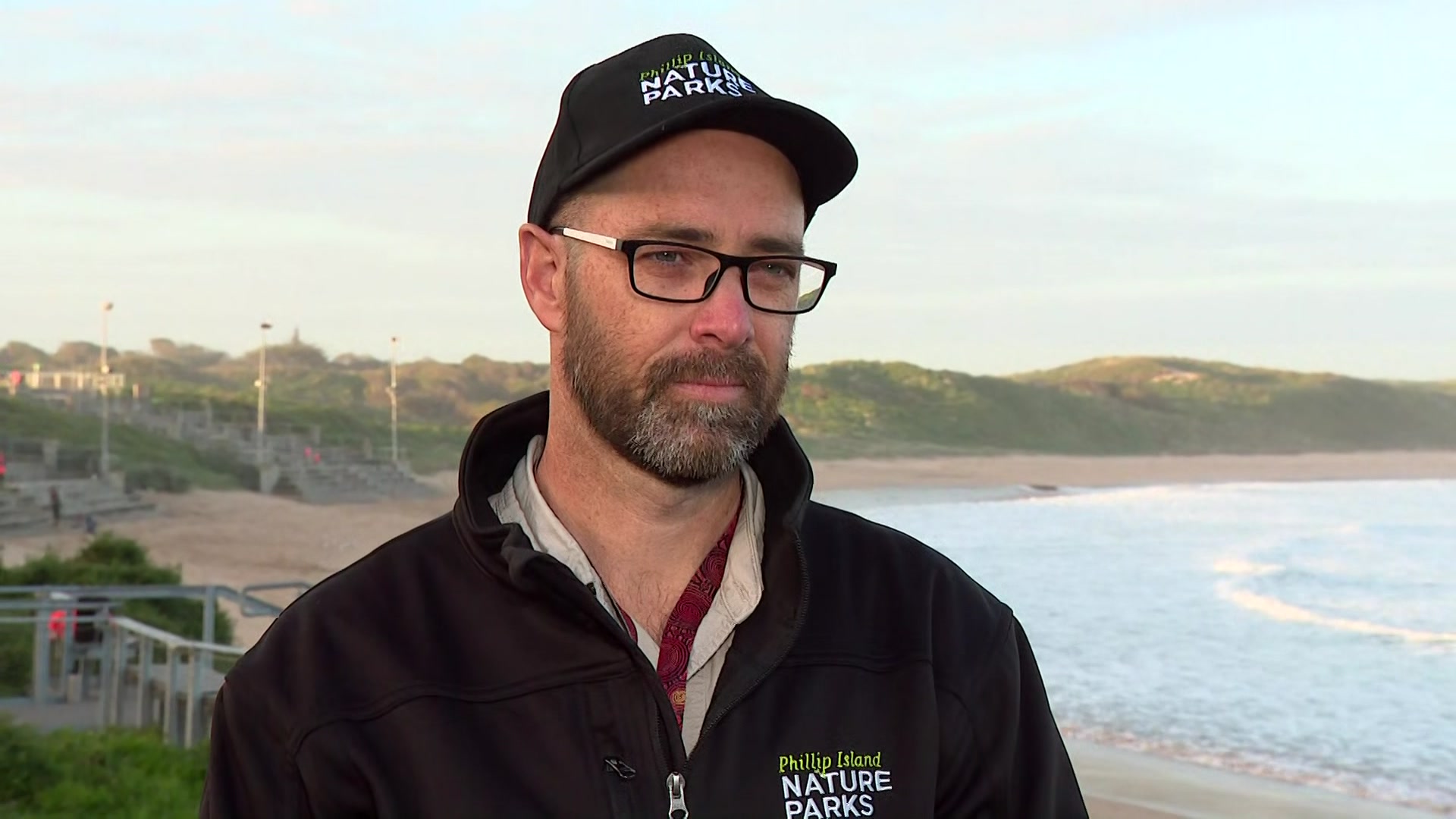 A bearded man wearing glasses, a black cap and black jumper stands on a beach.